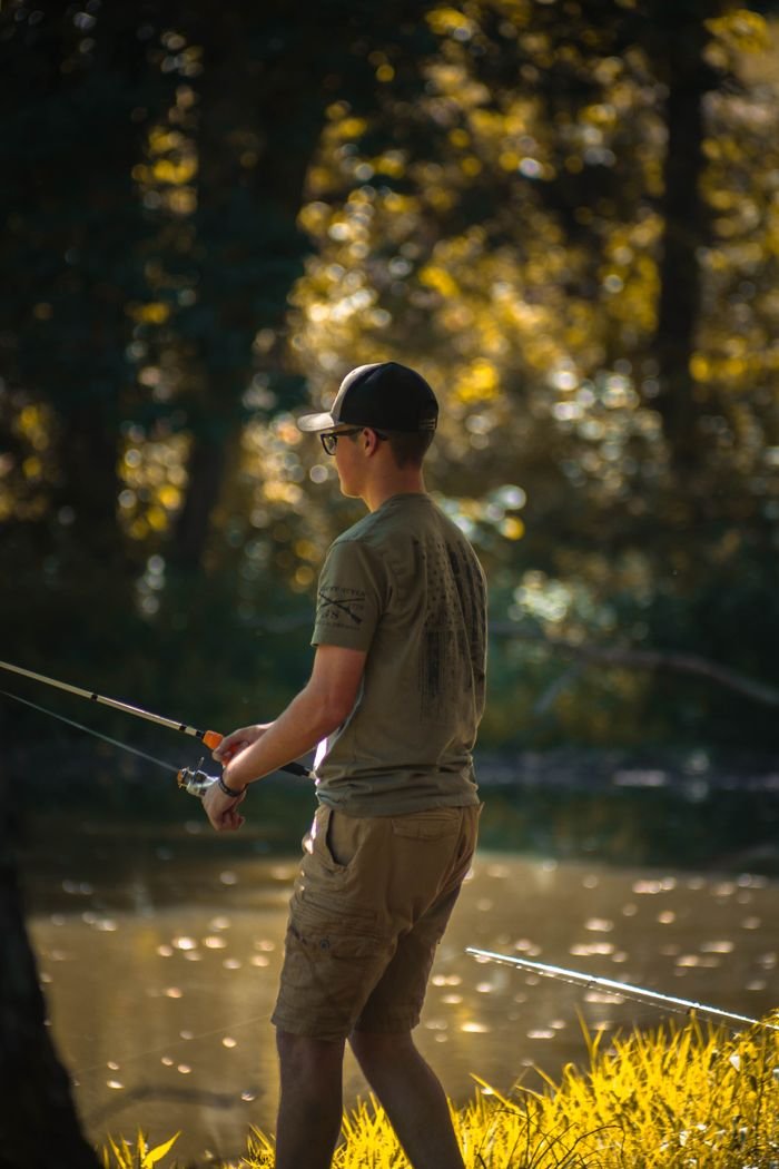 A young man stands on a riverbank fishing in a sunlit forest, casting his line.